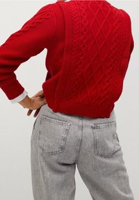 Roter gestrickter Pullover mit strukturierten Zopfmustern und gerippten Details, kombiniert mit hellgrauen, hoch taillierten Jeans mit Rückentaschen.