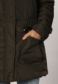 Veste parka rembourrée de couleur vert foncé, dotée d'une taille ajustable et de poches latérales, avec un tissu lisse et des détails cousus.
