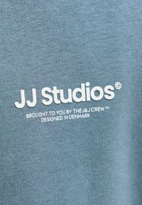 Sininen puuvillakangas, jossa on valkoista painettua tekstiä: "JJ Studios® Toimittanut J&J Crew™ Suunniteltu Tanskassa." Sileä rakenne.