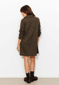 Robe chemise vert olive avec imprimé léopard, manches longues et poignets boutonnés, associée à des bottines marron foncé sur un fond uni.