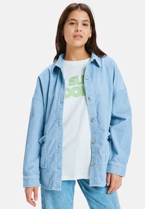 Giacca in velluto a coste azzurro chiaro con chiusura a bottoni, vestibilità oversize, due tasche laterali e texture a coste, indossata sopra una t-shirt bianca con grafica.