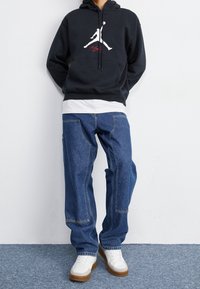 Felpa nera con logo Jumpman bianco, sopra una maglietta bianca, abbinata a jeans larghi blu e scarpe bianche con suola marrone.