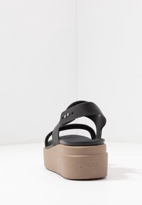 Sandales plates noires avec une texture lisse, des sangles larges et une semelle en caoutchouc beige. Présente un design minimaliste et des accents métalliques.