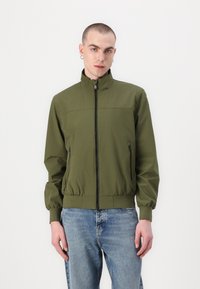 Maslinasto zeleni bomber jakna s visokim ovratnikom, prednjim zatvaračem i elastičnim manžetama. Ima dva bočna džepa sa zatvaračem i glatku, mat površinu.