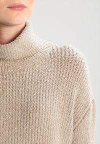 Beżowy sweter z golfem wykonany z dzianiny o prążkowanej strukturze. Miękka, teksturowana tkanina o luźnym kroju i lekkim połysku, podkreślająca pionowe linie.