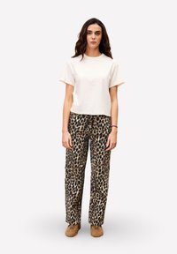 Femme se tenant de face, portant un t-shirt blanc uni, un pantalon à imprimé léopard et des chaussures marron, sur fond blanc.