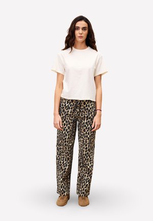 Femme se tenant de face, portant un t-shirt blanc uni, un pantalon à imprimé léopard et des chaussures marron, sur fond blanc.