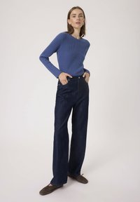 Blå ribbet striktrøje med langeærmer, parret med mørk denim vide bukser. Modellen står med hænderne på hofterne og har brune flade sko på.