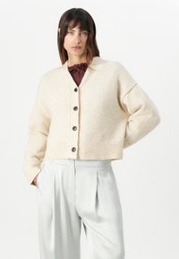 Beige kort cardigan i myk, lodden materiale med ribbet kant og store knapper. Kombinert med lyse, plisserte bukser.