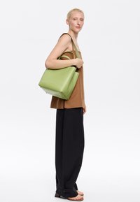 Green leather tote bag with structured design, dual top handles, and subtle gold branding. Worn with a brown top and black wide-leg pants.