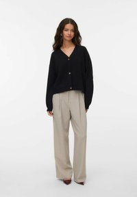 Kvinna med långt vågigt hår som bär en svart knappad cardigan, beige vida byxor och spetsiga bordeauxfärgade skor mot en vit bakgrund.