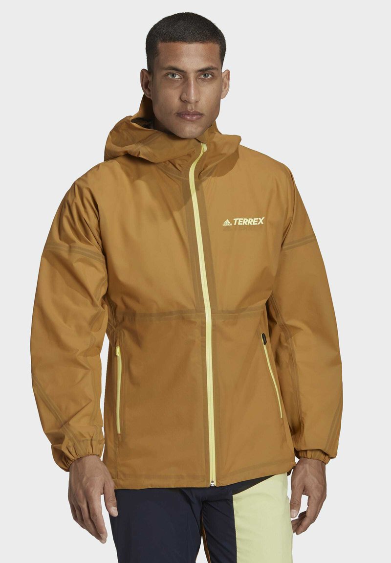 Chaqueta con capucha marrón y detalles en amarillo, que cuenta con cremallera completa y puños elásticos. El material parece ligero e impermeable.