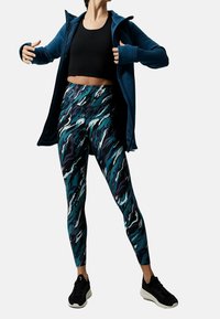 Dopasowany czarny top crop, zestawiony z legginsami w kolorze teal i granatowym wzorze w faliste linie. Przykryty niebieską kurtką zapinaną na zamek i czarnymi sneakersami.