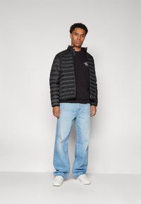 Giubbotto puffer nero con un design trapuntato leggero, indossato sopra una maglietta nera, abbinato a jeans baggy blu chiaro e sneakers bianche.