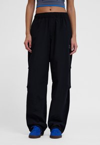 Zwarte cargobroek met een relaxte fit, elastische tailleband en zijzakken. G gedragen met blauwe suède schoenen. Glad stofoppervlak.