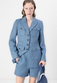 Kék tweed blézer, két elülső zsebbel és fekete gombokkal, hozzá illő, rojtos szélű sorttal párosítva. Sima textúra, szabott fazon.