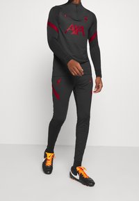 Sort atletisk træningsdragt med røde accenter, der inkluderer en lynlås-langærmet top og matchende bukser. Nike og Liverpool logoer er synlige.