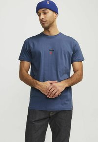 Tricou din bumbac albastru, cu mâneci scurte, având inscripția "R.D.D." și un cerc roșu. Este asortat cu blugi negri din denim și purtat cu o căciulă din tricot albastră.