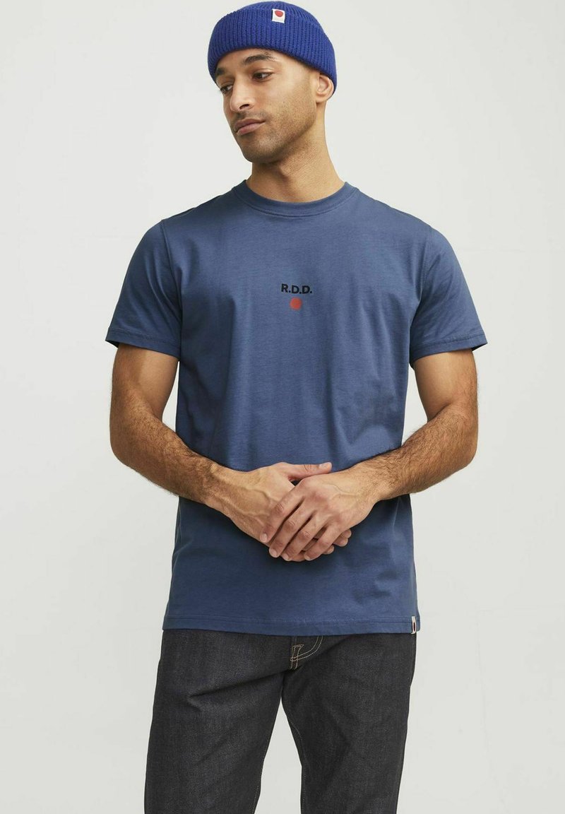Tricou din bumbac albastru, cu mâneci scurte, având inscripția "R.D.D." și un cerc roșu. Este asortat cu blugi negri din denim și purtat cu o căciulă din tricot albastră.