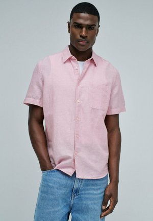 Joven con el cabello corto, vistiendo una camisa de manga corta de color rosa claro y vaqueros azules, de pie con una mano en el bolsillo.