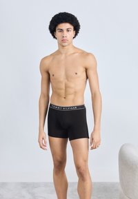 Μαύρο ανδρικό εσώρουχο τύπου μποξέρ με λευκή μέση που φέρει το λογότυπο "TOMMY HILFIGER", εφαρμοστό, παρουσιασμένο σε μοντέλο με αθλητική σωματοδομή σε ουδέτερο φόντο.