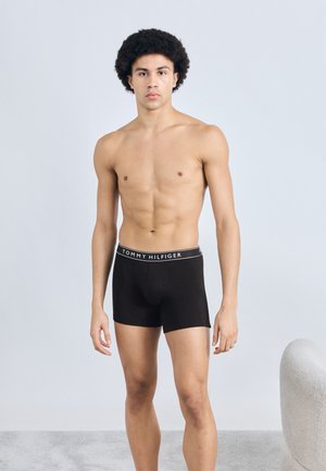 Shorts boxer noirs avec une ceinture blanche portant le logo "TOMMY HILFIGER", ajustés, présentés sur un mannequin athlétique devant un fond neutre.