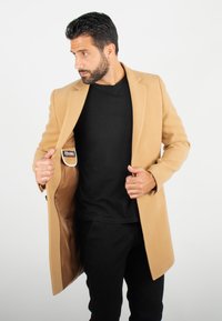 Manteau en laine beige avec un col cranté, une fermeture à un bouton à l'avant, et une poche intérieure. Porté par-dessus une chemise noire et un pantalon noir.