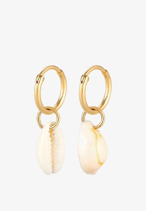 Boucles d'oreilles en or avec des coquillages de vache blancs. Les coquillages présentent des textures lisses et des motifs naturels, suspendus à un anneau doré.