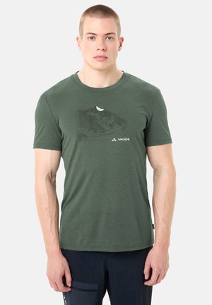 Homme portant un t-shirt vert olive avec un motif de paysage montagneux noir et le texte "Vaude", associé à un pantalon noir sur un fond uni.