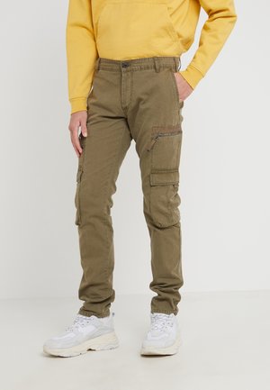 Person iført olivengrønne cargobukser med lynlås- og klaplommer, en gul hættetrøje og hvide chunky sneakers, stående mod en lys baggrund.