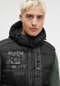 Schwarze gepolsterte Jacke mit hohem Kragen, versehen mit gestickten Logos, Textdetails und einem vertikalen Riemen mit der Aufschrift "BLUE CREW."