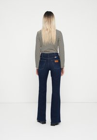 Ausgestellte blaue Jeans aus Denim mit dunkler Waschung, die Taschen und ein braunes Lederpatch aufweisen, kombiniert mit einem schwarz-weißen gestreiften Langarmshirt.