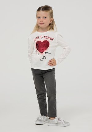 Camicia bianca a maniche lunghe con un design a cuore rosso in paillettes e testo stampato, abbinata a pantaloni grigi e sneakers argentate.