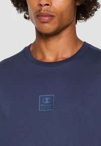 Mørkeblå bomuld t-shirt med rund halsudskæring, der har et lille lyseblåt Champion-logo på brystområdet. Glat tekstur.