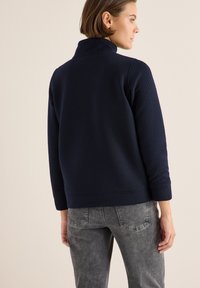 Marinblå, strukturerad fleece-pullovar med hög krage, som har en avslappnad passform och ribbade ärmslut. Kombinerad med grå, utslitna jeans.