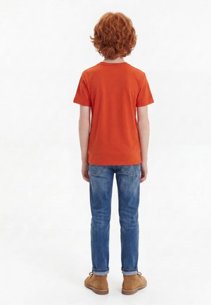 Dziecko o kręconych rudych włosach, ubrane w pomarańczowy T-shirt, niebieskie dżinsy i beżowe buty, stoi tyłem na jednolitym białym tle.