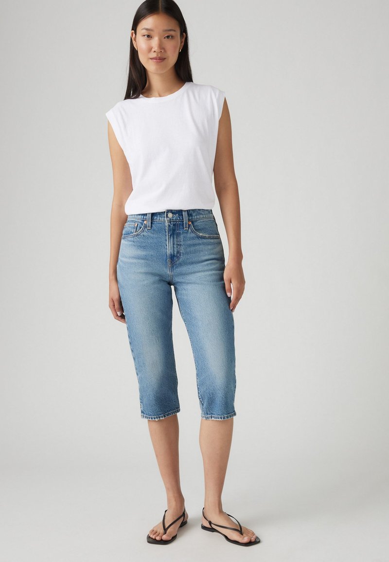 Haut sans manches blanc associé à un jean cropped bleu clair, à taille haute et avec un ourlet effiloché. Porte des tongs noires.