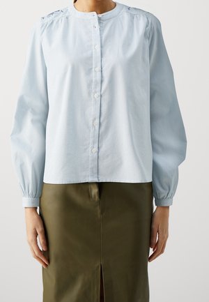 Camicia button-up azzurro chiaro con righe verticali, scollo rotondo e maniche a sbuffo. Indossata con una gonna di pelle verde con uno spacco.