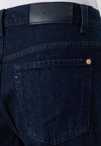 Dunkelblaue Denim-Jeans mit einer glatten Textur, hinterer Tasche mit goldenen Hardware-Details und einem strukturierten schwarzen Lederetikett am Bund.