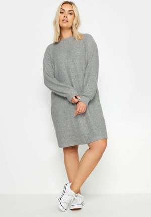 Graues Strickkleid mit geribbelter Textur, langen Ärmeln und rundem Ausschnitt, kombiniert mit weißen Sneakers. Das Kleid hat eine entspannte Passform und eine mittellange Länge.