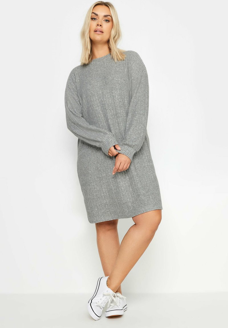Abito maglione grigio con texture a costine, maniche lunghe e scollo rotondo, abbinato a sneakers bianche, presenta una vestibilità rilassata e un taglio a lunghezza media.