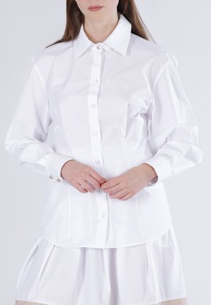 Kvinde iført en hvid skjorte med lange ærmer og krave samt et hvidt skørt, hænderne foldet ved taljen.