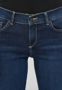 Dark blue denim jeans feature a slim fit, metal button closure, and subtle fading at the seams, with visible stitching accents on the pockets.