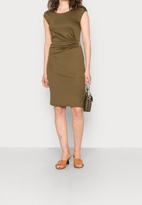 Olijfgroene jurk met korte kapmouwen en een twistdetail aan de zijkant. Model houdt een kleine olijfgroene tas vast en draagt bruine slidesandalen.