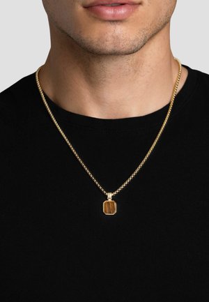 Halskette aus goldfarbenem Metall mit einem sechseckigen Anhänger und braunem Stein, getragen über einem schwarzen T-Shirt.