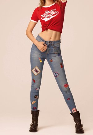 Frau trägt ein rotes, gebundenes Croptop und blaue Jeans, die mit bunten Aufnähern verziert sind, kombiniert mit schwarzen Stiefeletten mit Stern-Details.