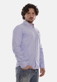 Camisa de botonadura en azul claro hecha de algodón, con mangas largas, un cuello y un pequeño logo en el pecho. Combinada con jeans desgastados.