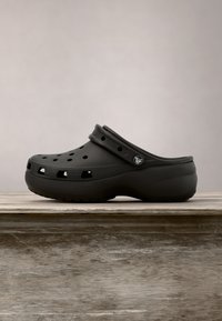 Sabot noir à enfiler avec des trous de ventilation et une bride réglable au talon, présenté sur une surface en bois avec un fond uni.