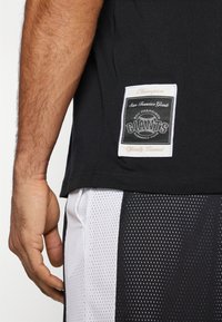 Svart bomull t-skjorte med et vevd merke som har "San Francisco Giants" i hvitt og svart, parret med hvite og svarte mesh-shorts.