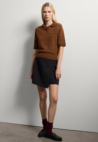 Maglione polo in misto lana a maniche corte marrone abbinato a una gonna a portafoglio nera elegante. Modelli scarpe nere e calzini bordeaux. Sfondo minimalista.